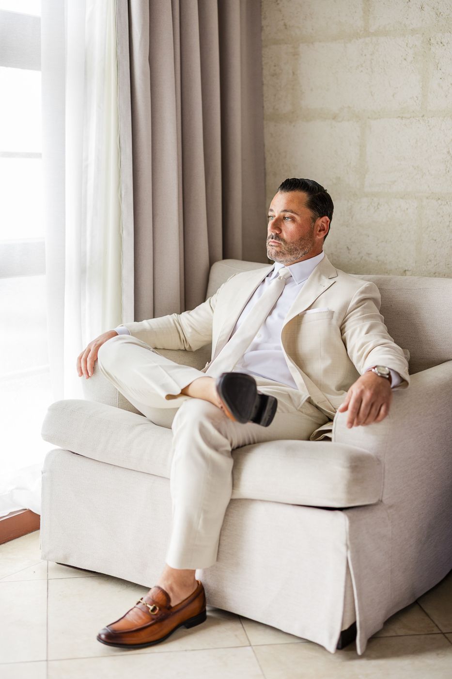 A well-dressed man in a beige suit relaxes in a white armchair, gazing thoughtfully toward a bright window.