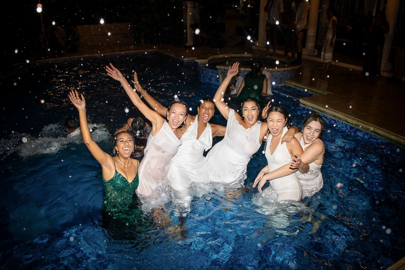 A group of people splashing and playing in a swimming pool at night with illuminated water.