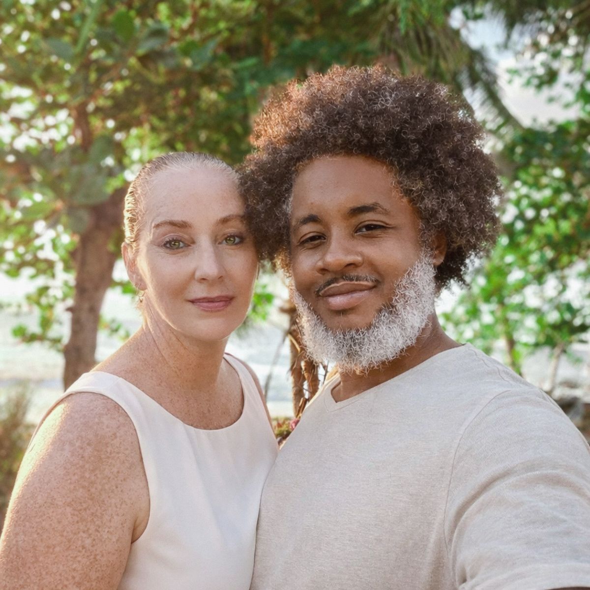 Wedding couple in white outfits posing together outdoors under trees.