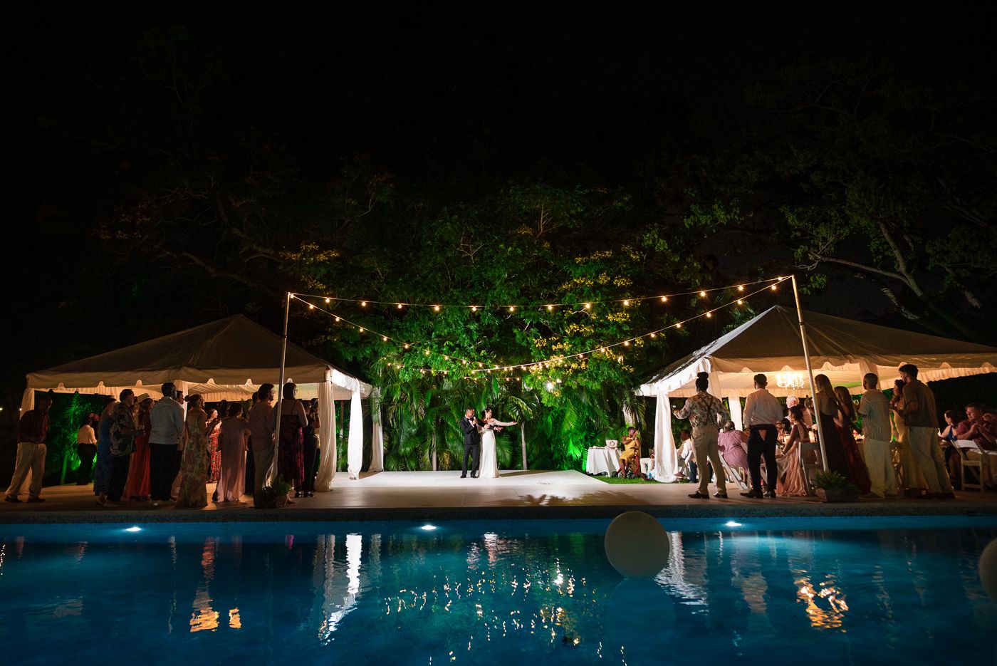 String lights illuminate an outdoor wedding reception venue at night with lush greenery and white tents.