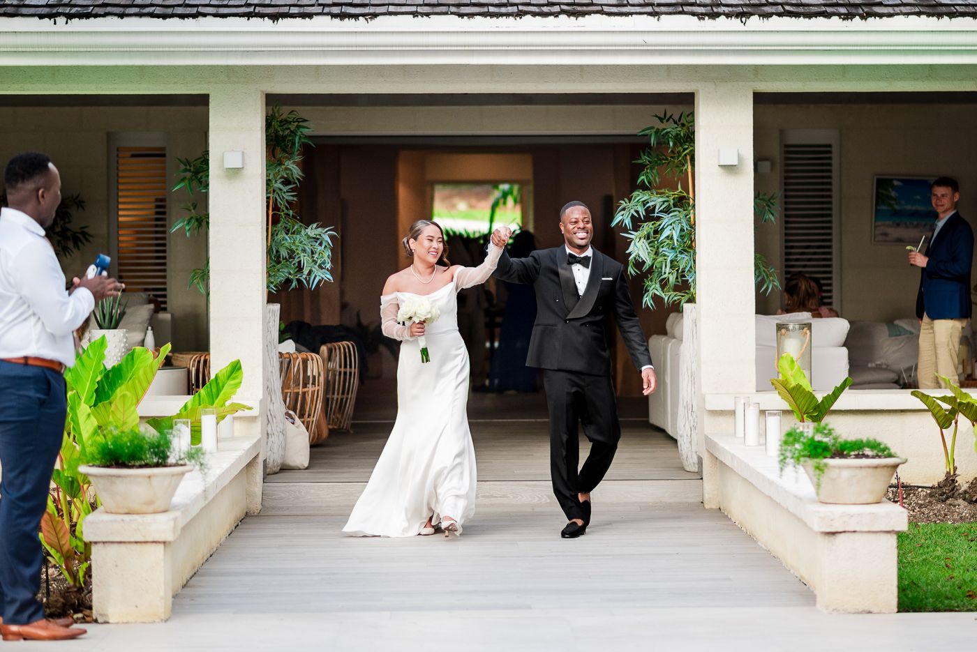 Wedding guests walk along a covered outdoor walkway with white columns during an elegant garden ceremony.