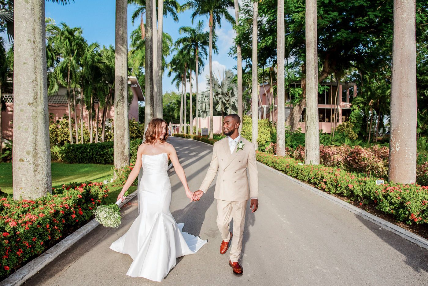Couple walks hand in hand down palm tree lined pathway in tropical wedding venue.