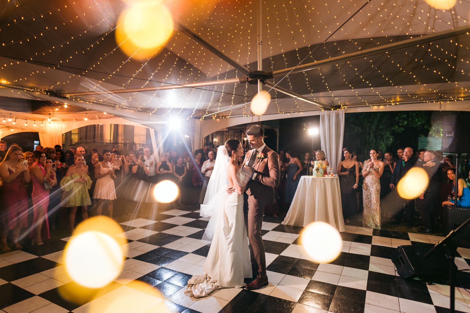 A couple shares their first dance on a black and white checkered floor under glowing lights at their wedding reception.