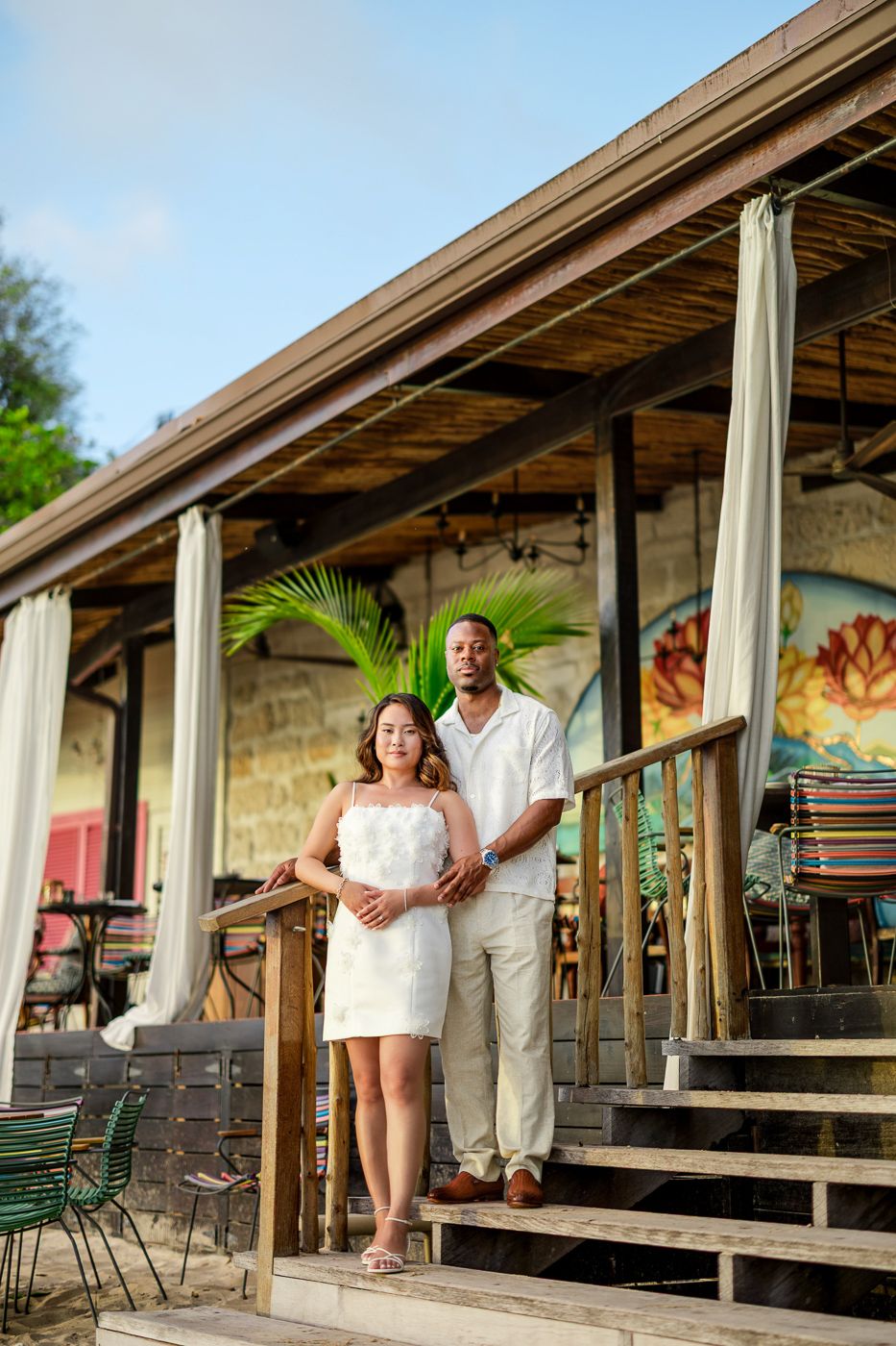 A couple poses on wooden steps of a tropical restaurant with palm decor.