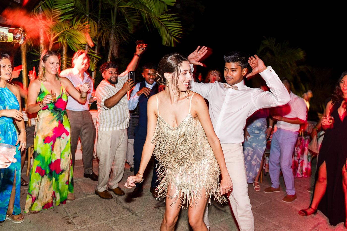 Large group of party guests dancing outdoors under colorful lighting with palm trees.