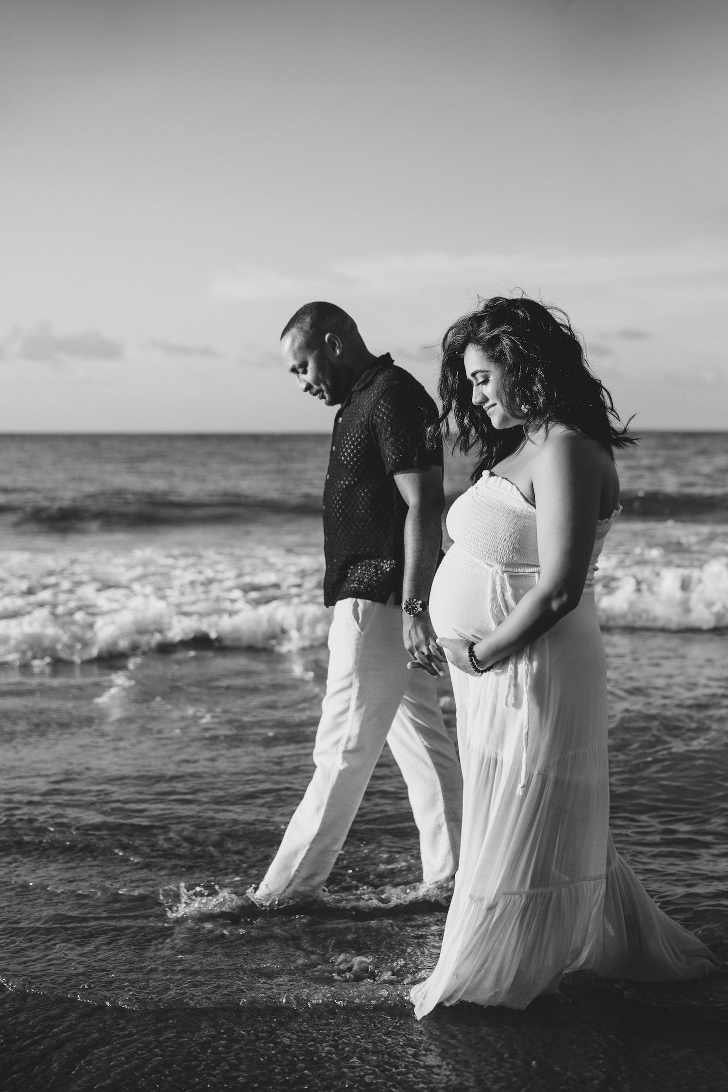 Black and white photo of a pregnant couple walking along the ocean shore, woman in flowing white dress holding her baby bump.