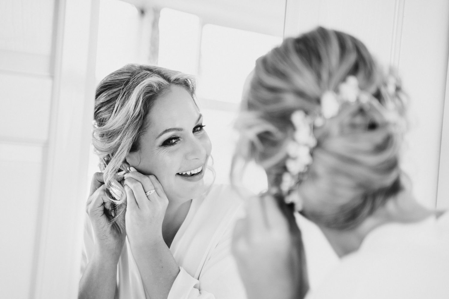 Bride smiling in mirror while putting on earrings, black and white wedding photo.
