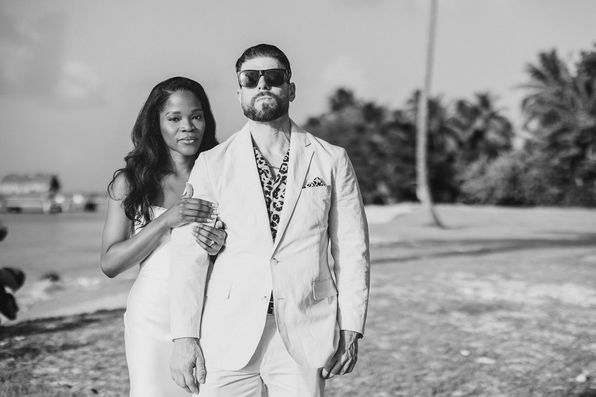 Black and white photo of an elegant couple in white outfits posing on a tropical beach with palm trees.