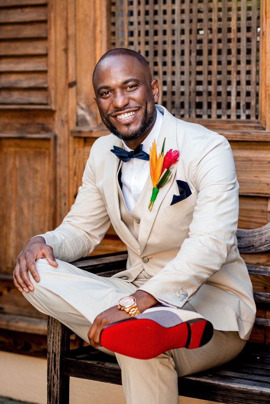 Smiling groom in cream suit with colorful boutonniere and red socks sits on wooden bench.