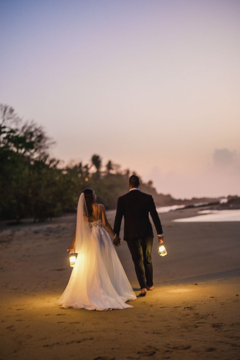 Newlywed couple walks along beach at sunset holding lanterns casting warm glow on sand.