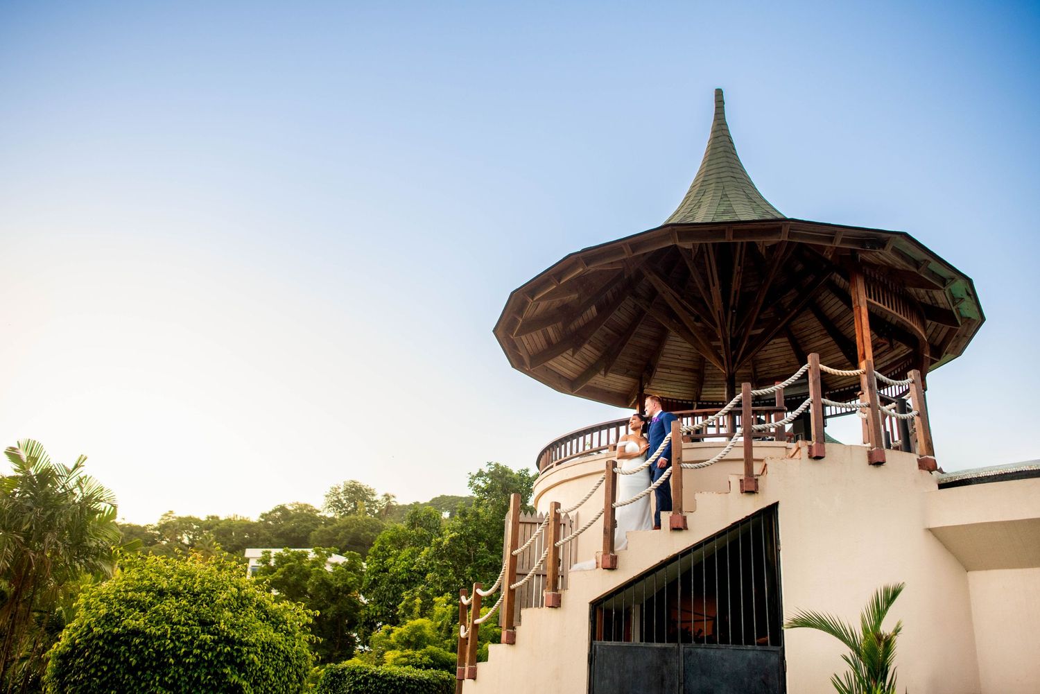 Couple stands on elegant gazebo with pointed thatched roof overlooking lush tropical gardens at sunset.