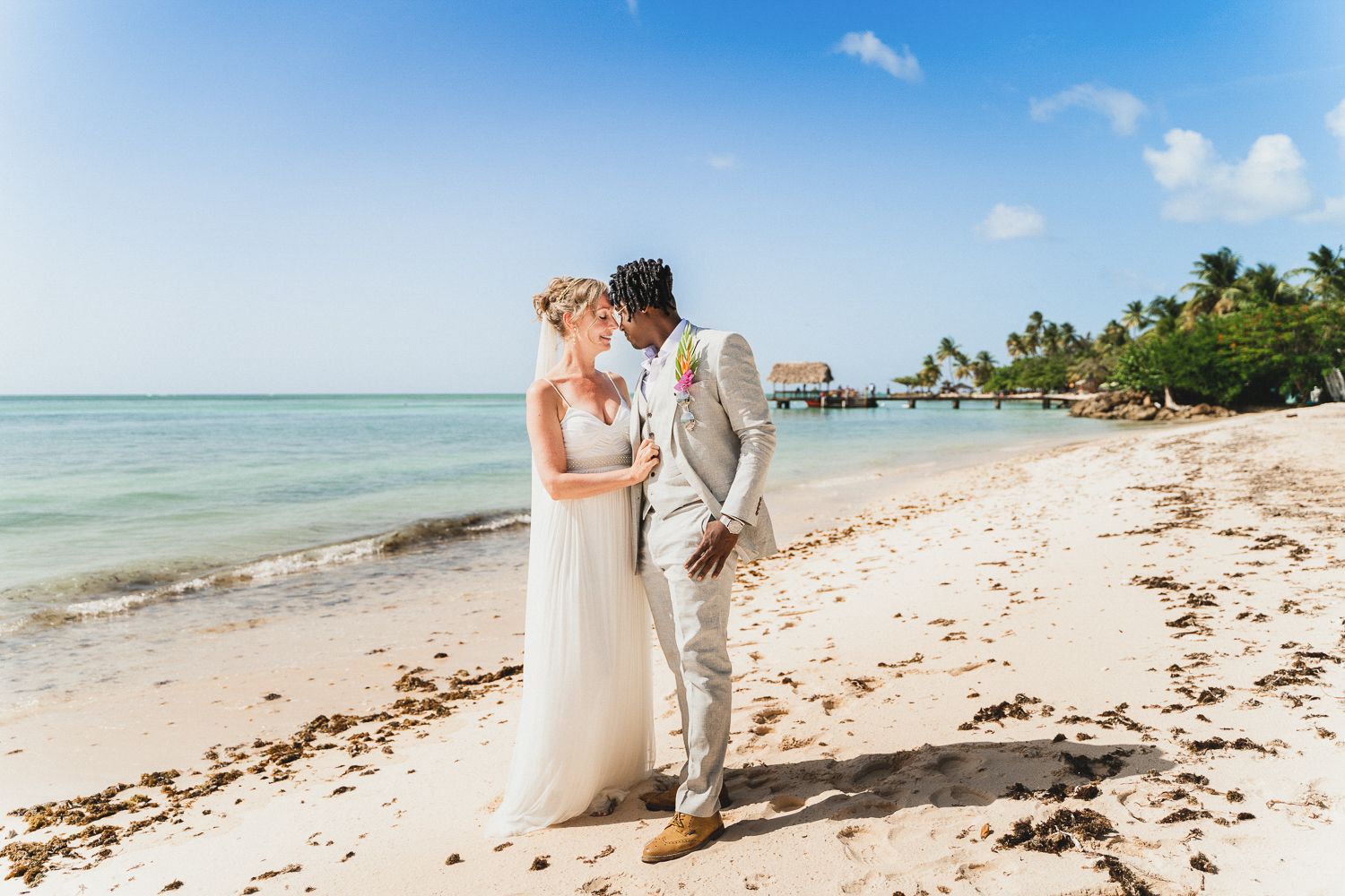 Couple in wedding attire embracing on a tropical beach with turquoise water and palm trees in the background.