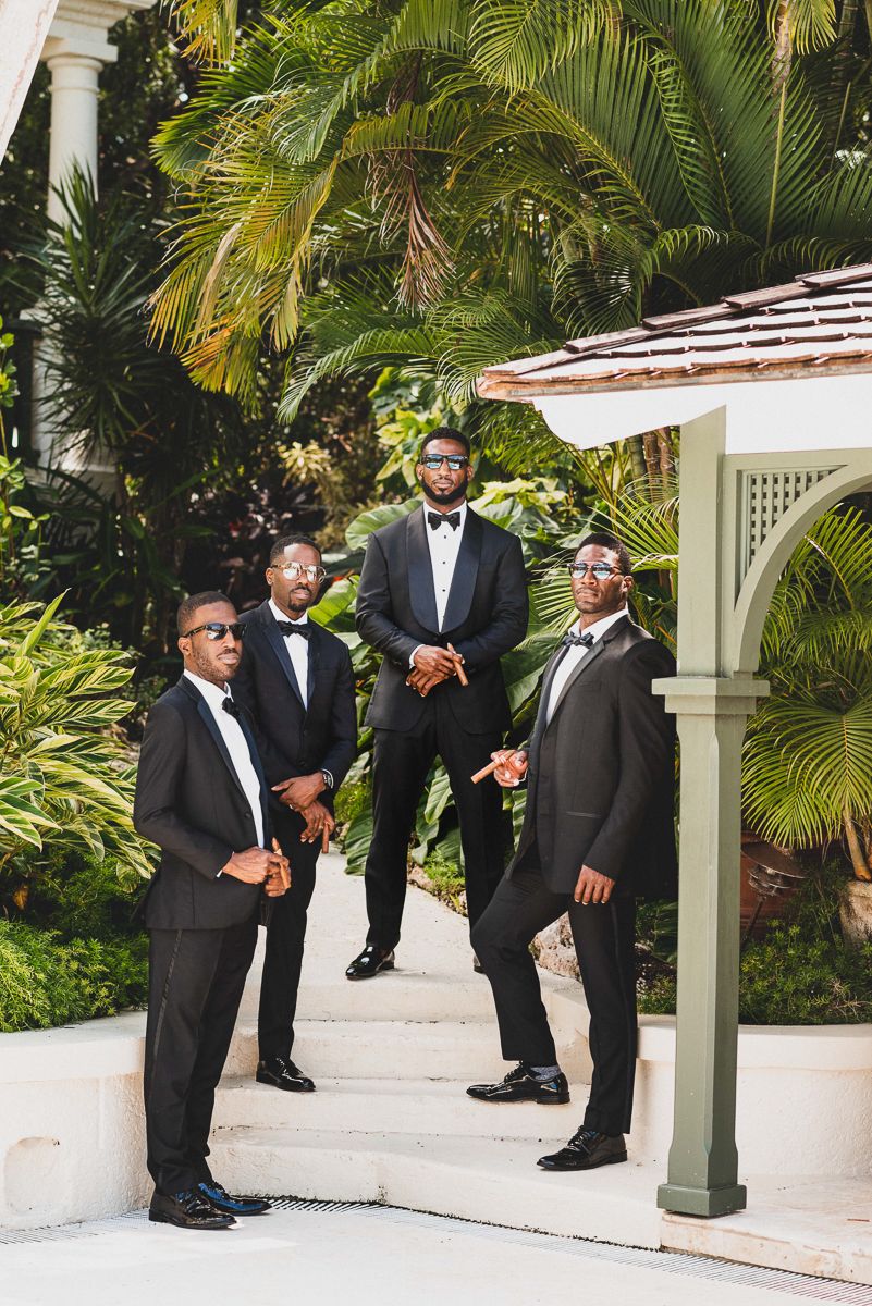 Four groomsmen in black tuxedos and bow ties pose outdoors near tropical palm trees and a white gazebo.