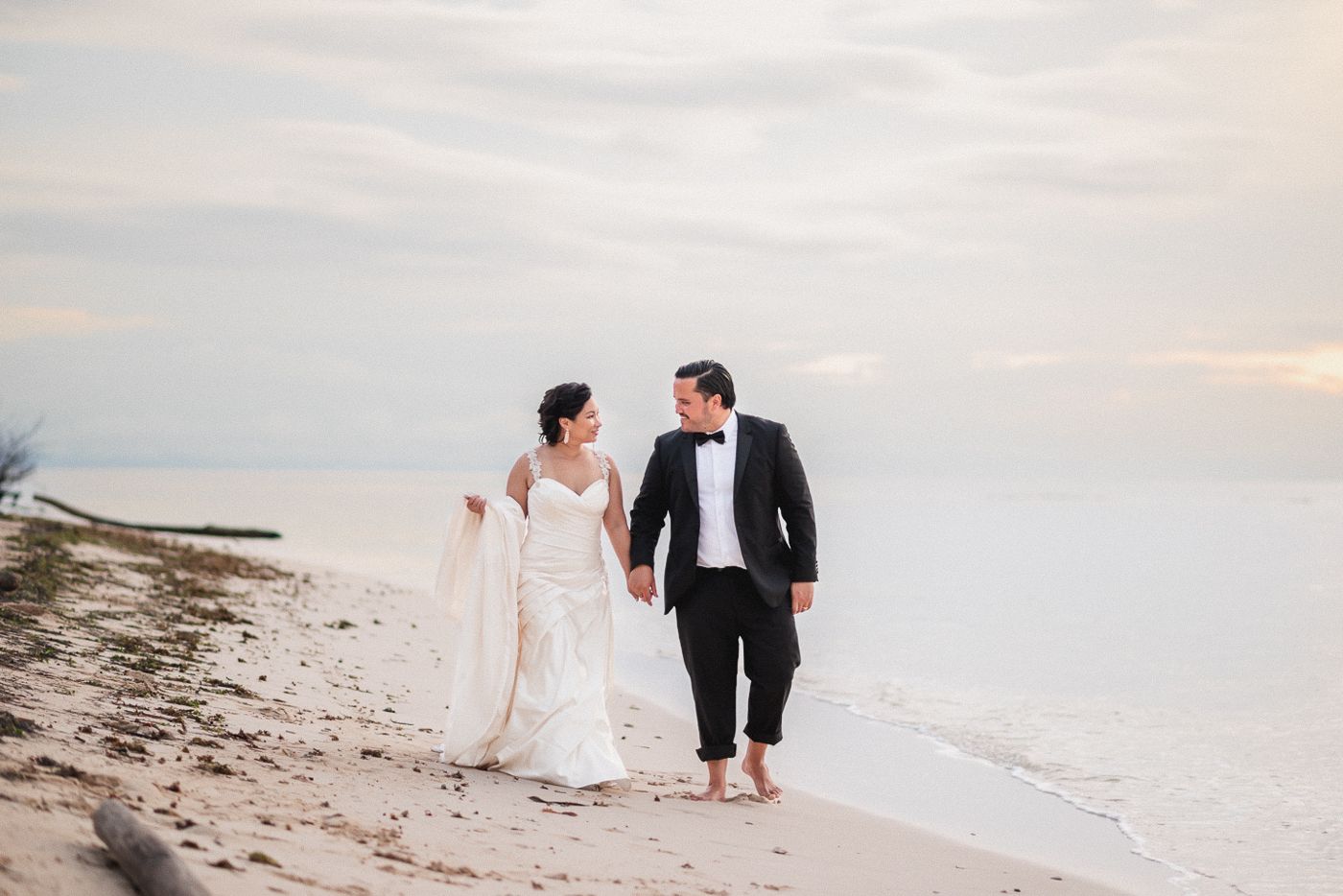 Bride and groom walk hand-in-hand along a serene beach at sunset in wedding attire.