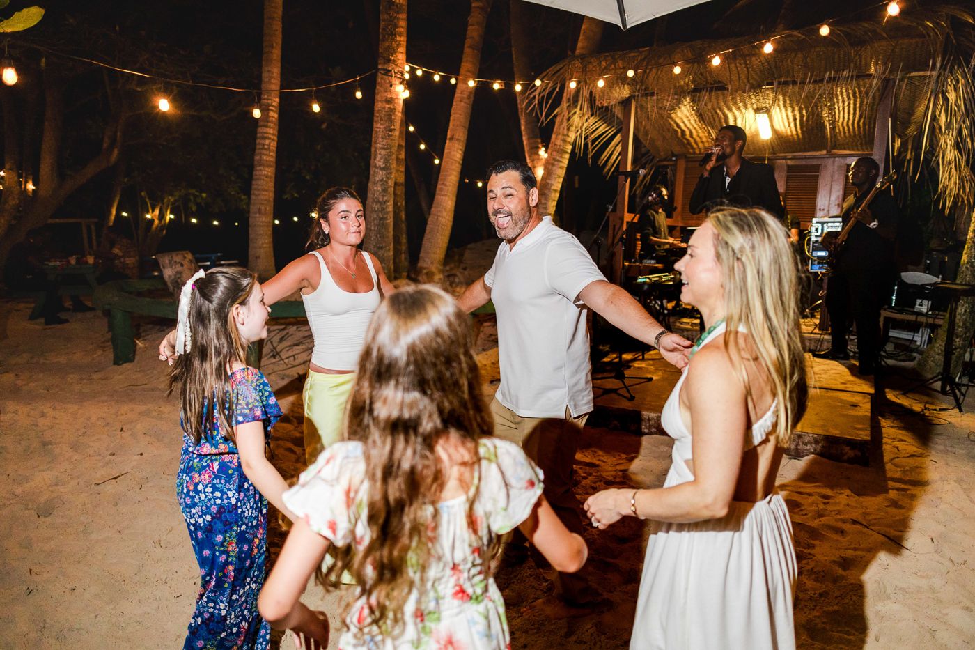 Group of adults and children dancing together at outdoor tropical beach party under string lights at night.