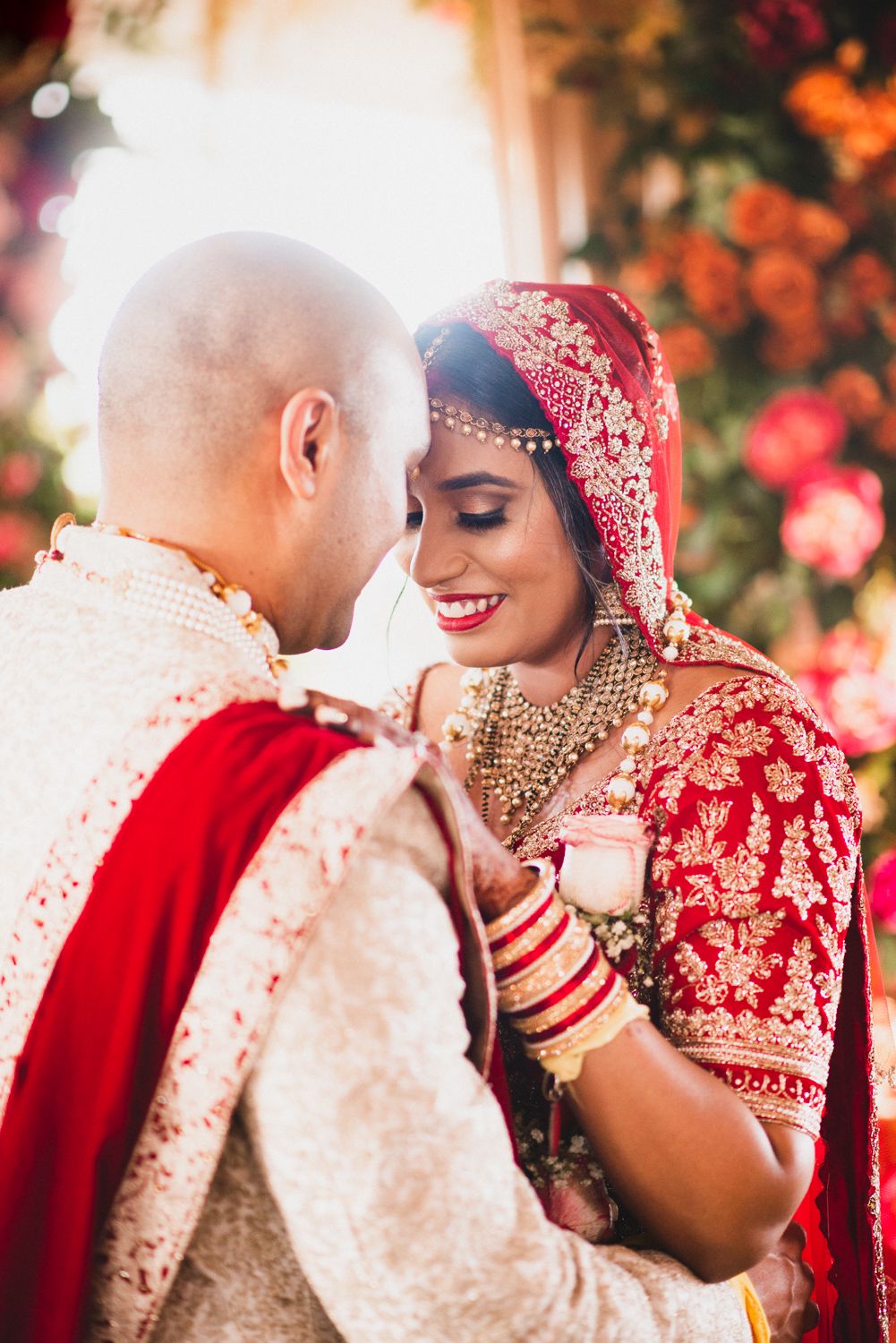 Indian bride in red lehenga and groom in sherwani share an intimate moment at traditional Hindu wedding ceremony.