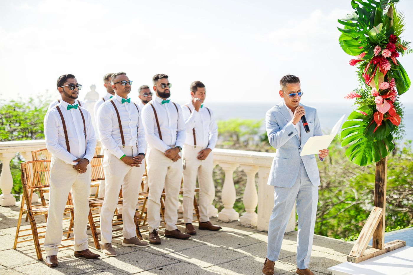 Groomsmen in light colored suits and suspenders stand at attention during outdoor ceremony.