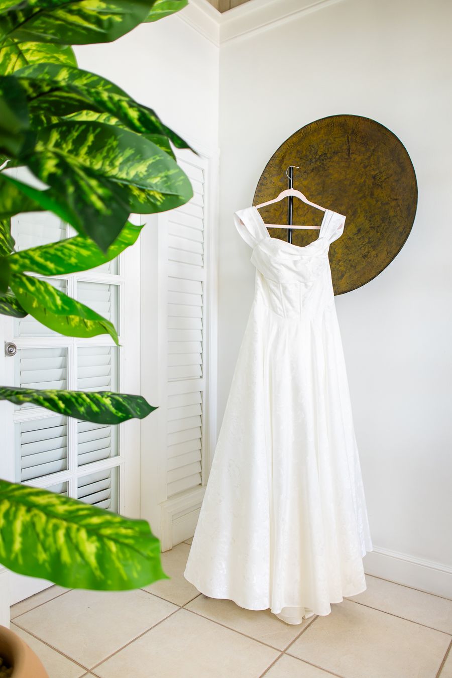 White wedding dress displayed in a bright room with tropical green leaves in the foreground.