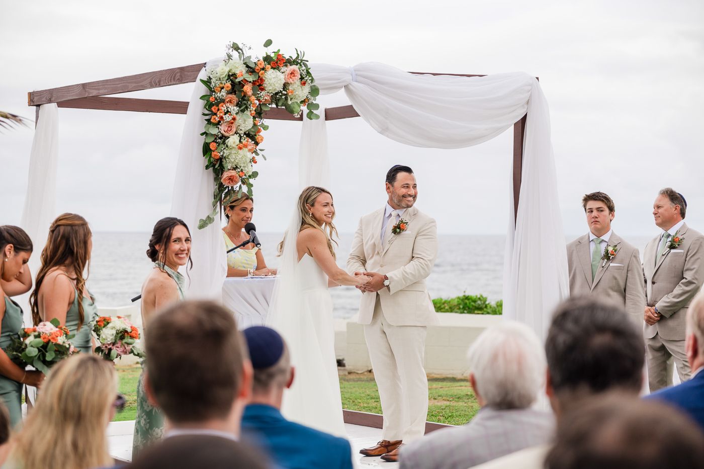 Couple exchanges vows at outdoor beach wedding ceremony under floral arch with white draping and ocean backdrop.