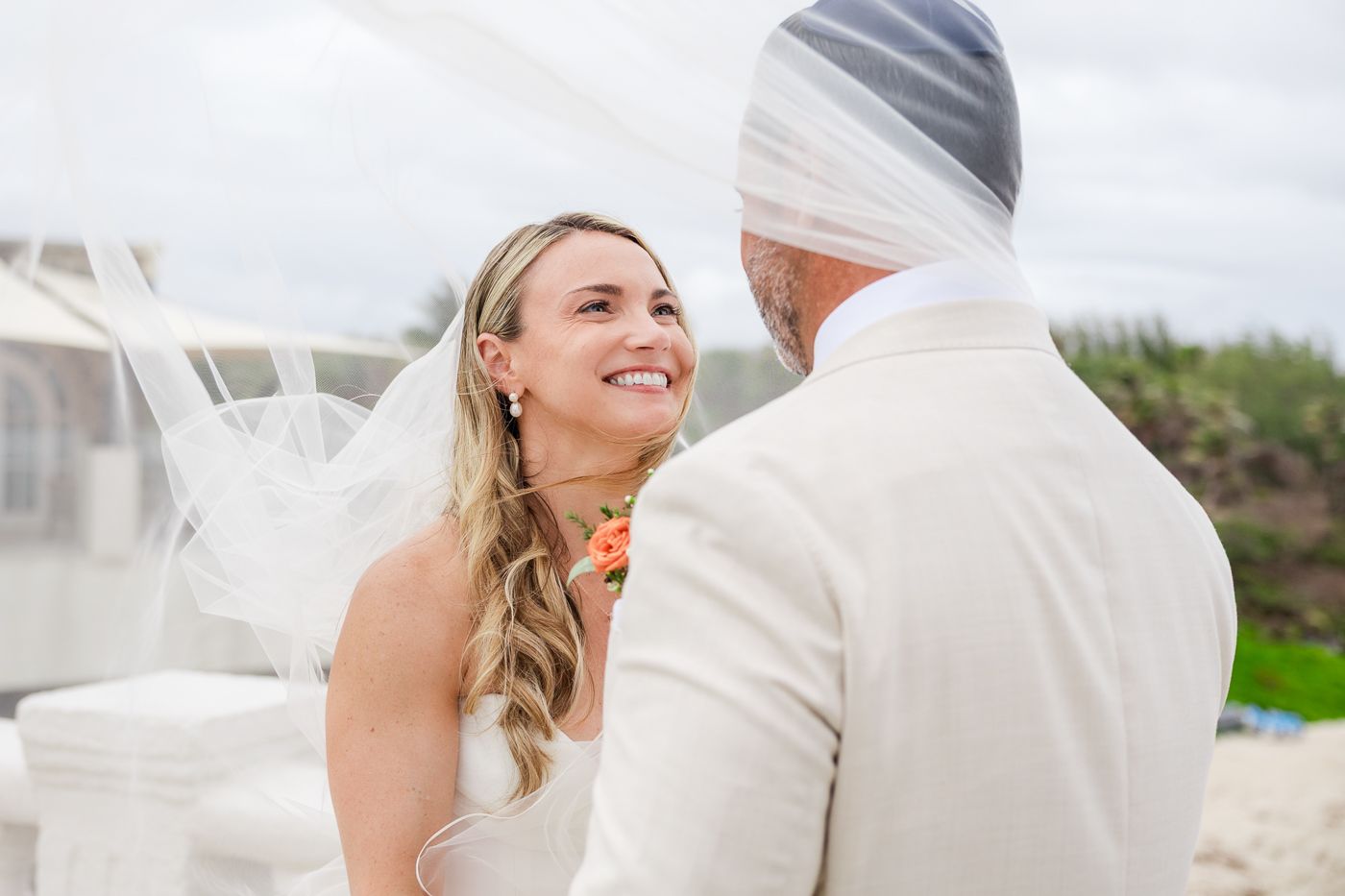 Bride smiling at groom during outdoor beach wedding ceremony, wearing white dress with veil blowing in the wind.