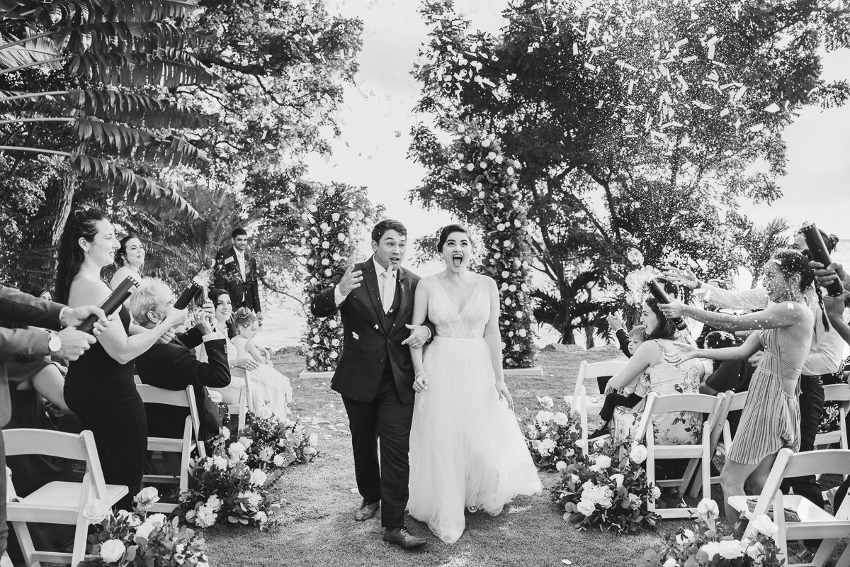 Black and white photo of a joyful bride and groom walking down the aisle at an outdoor wedding ceremony.