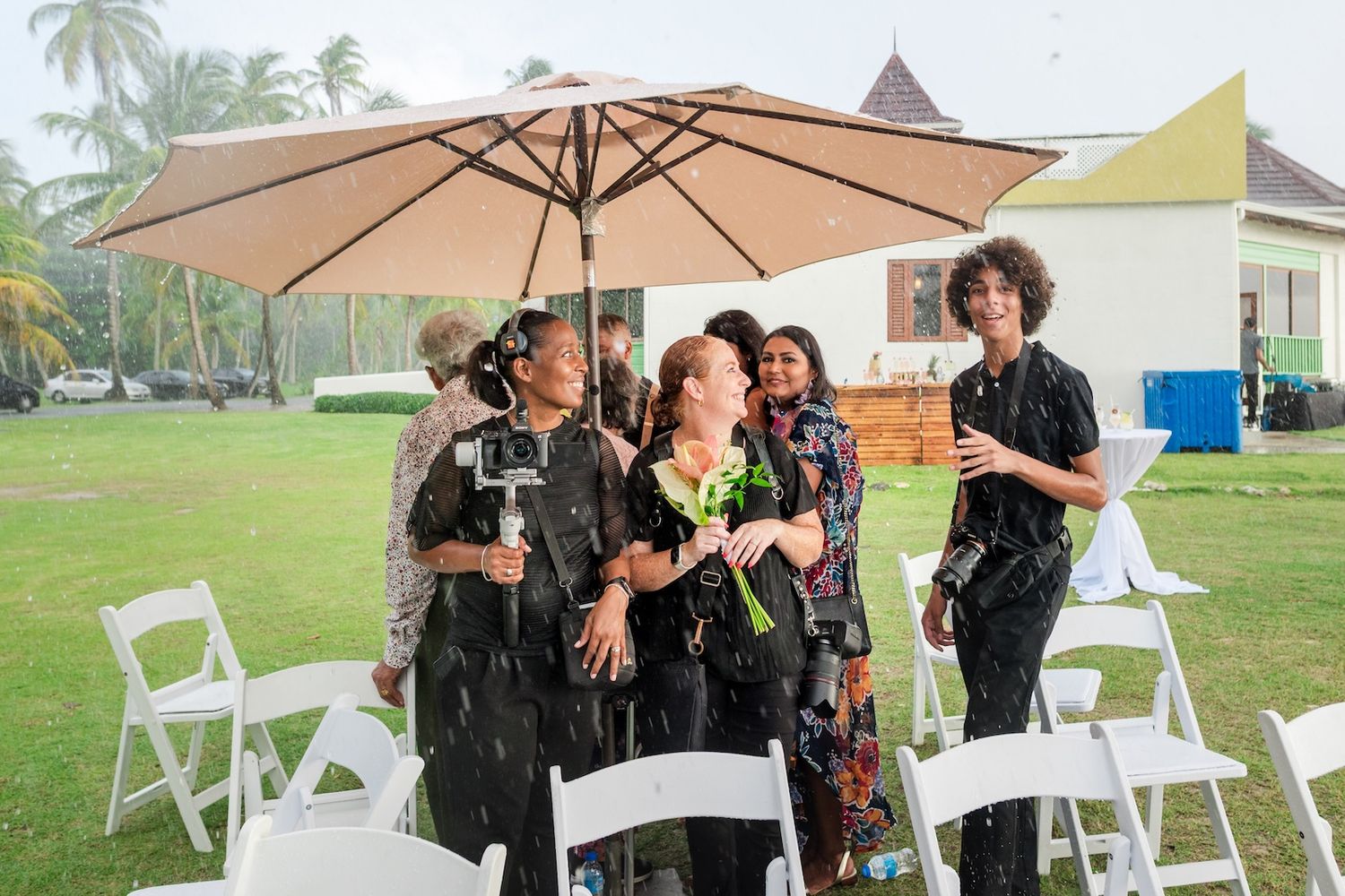 Women laughing under umbrella at outdoor wedding ceremony in rain, white chairs and tropical trees in background.