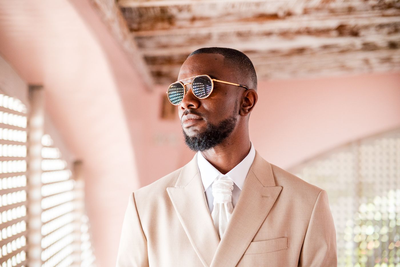 Someone in a beige suit posing in a hallway with latticed windows.