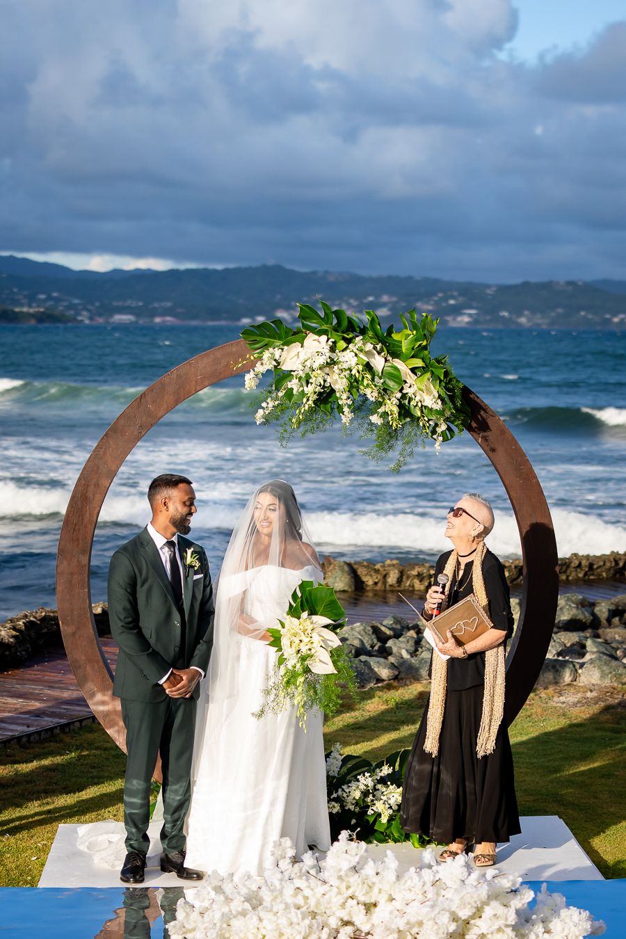 Oceanfront wedding ceremony under circular arch with floral decor and crashing waves in background.