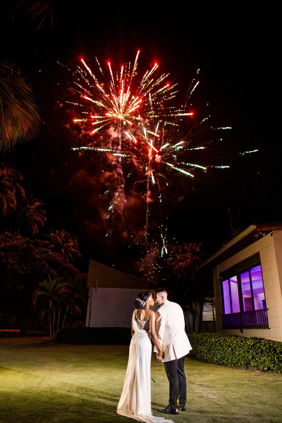 Wedding couple embraces while fireworks illuminate the night sky above.
