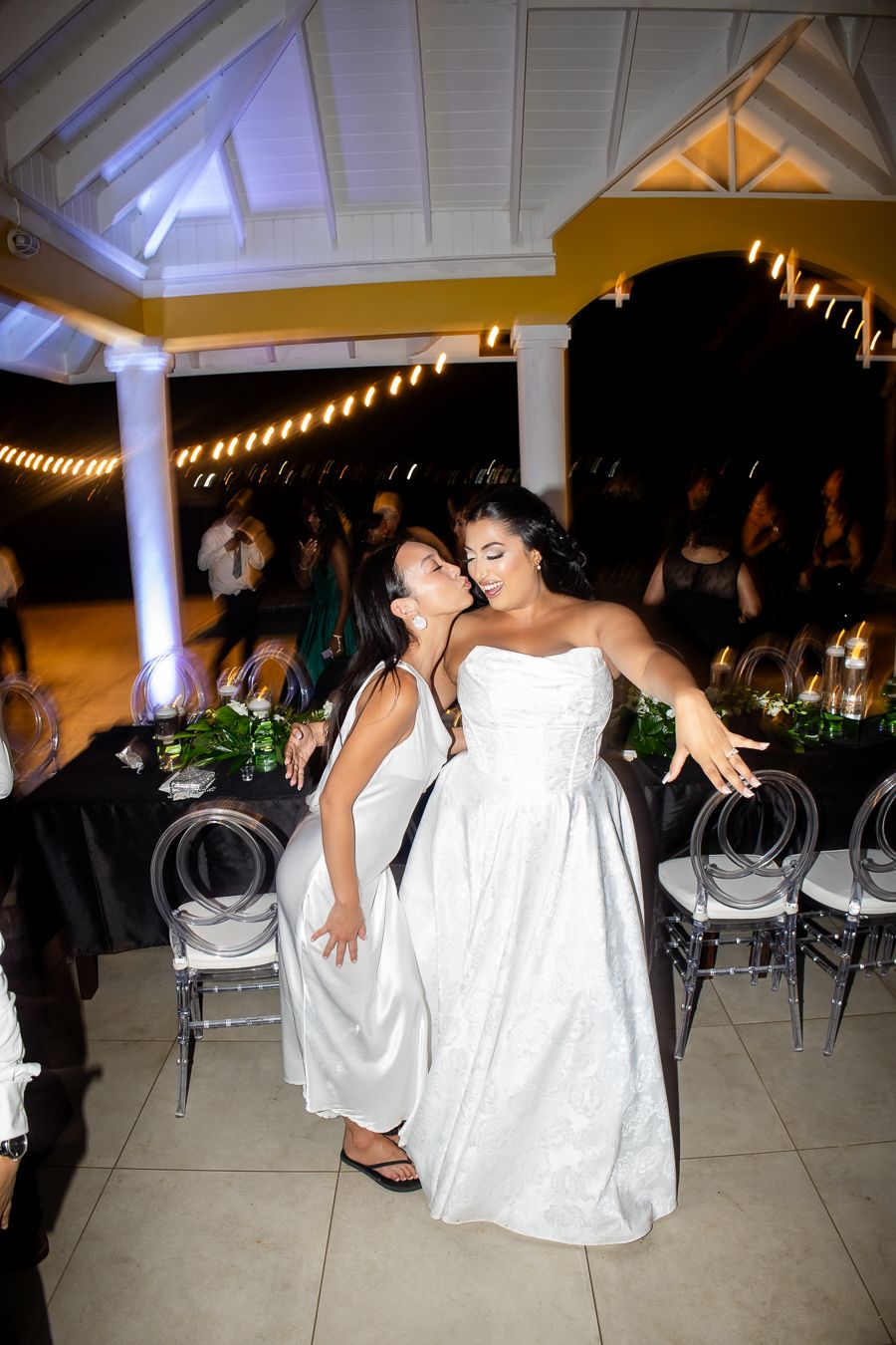Two people in white dresses dancing joyfully under string lights at wedding reception.