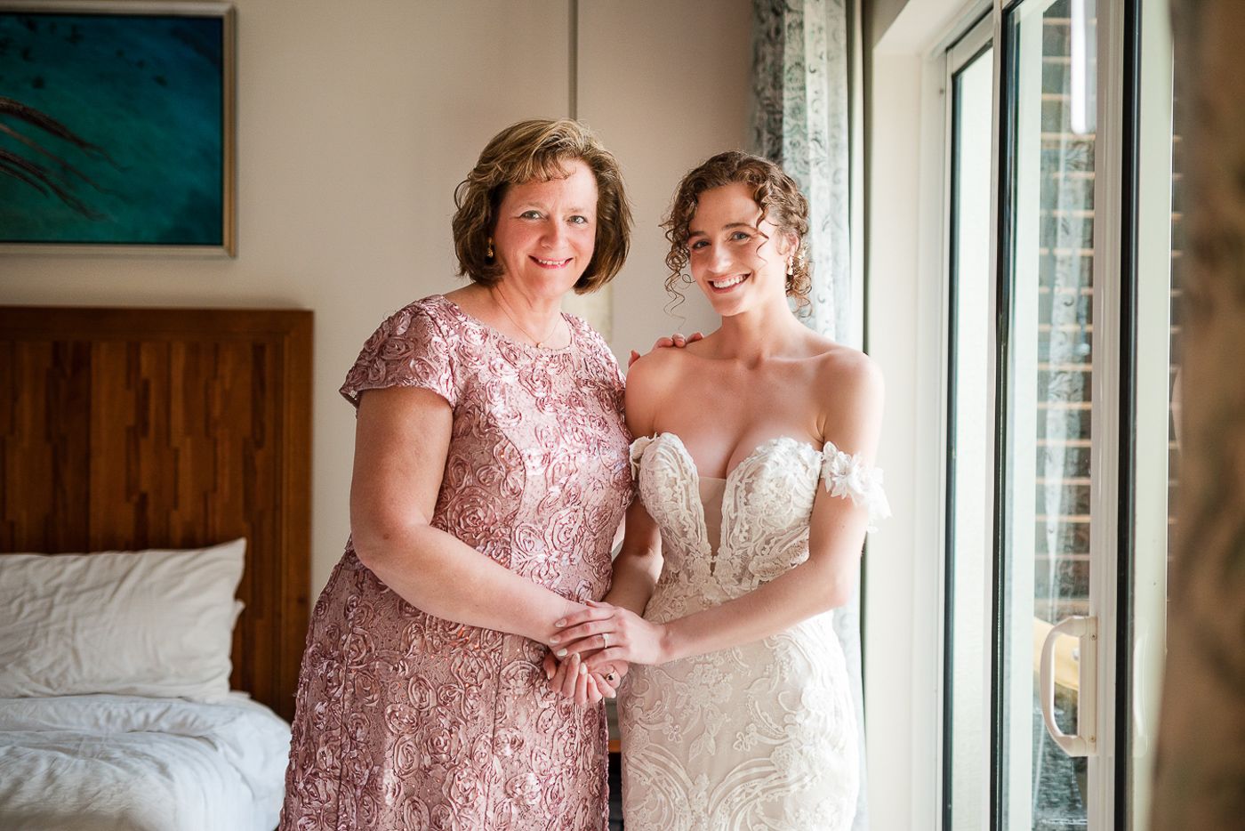 Two women in formal dresses share a tender moment by a window in an elegantly decorated room.