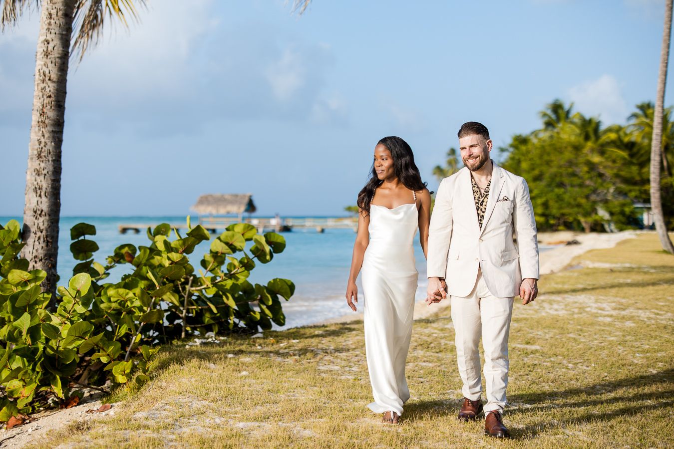 Couple in wedding attire walks hand-in-hand on tropical beach with palm trees and ocean views.