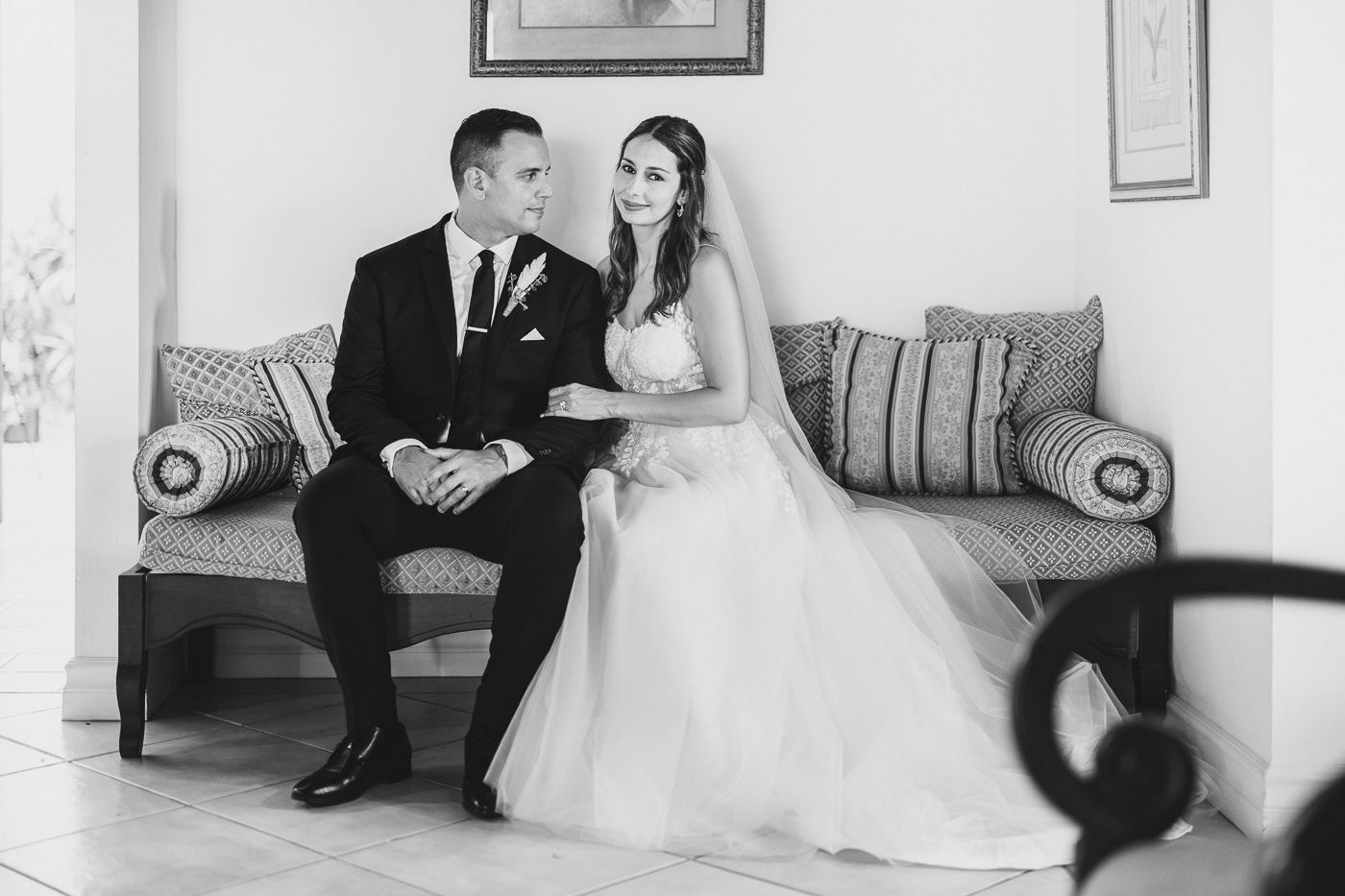 Black and white photo of a bride and groom sitting together on an elegant sofa, sharing a tender moment on their wedding day.