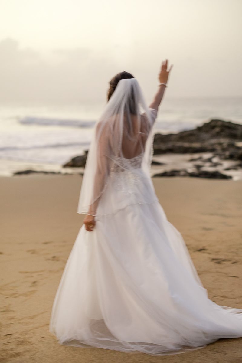 Bride in white gown and veil stands on sandy beach, arm raised, with ocean waves and rocks in background.