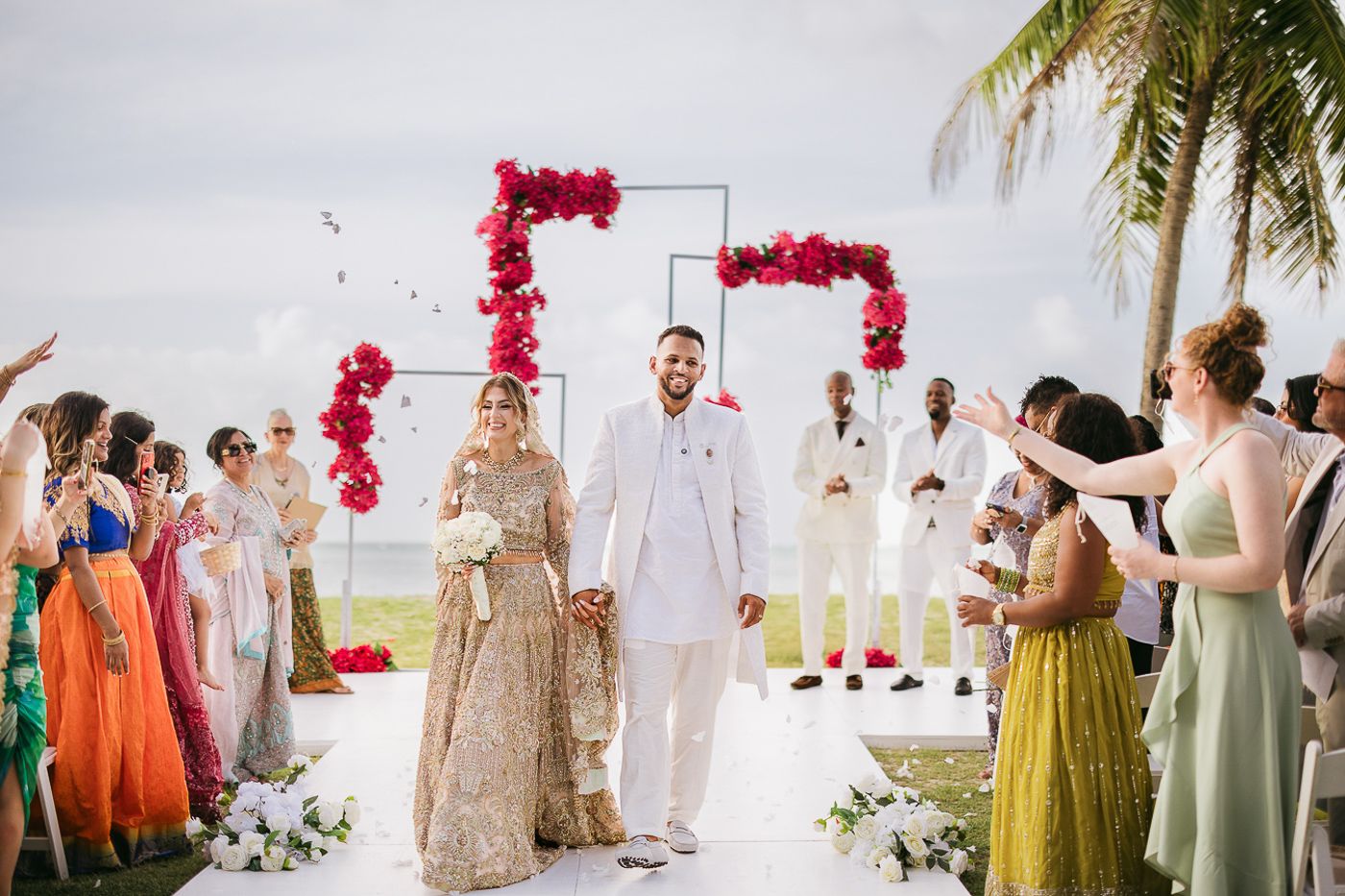 Couple walks down aisle at outdoor Indian wedding ceremony with red floral arch, guests celebrating in colorful attire.