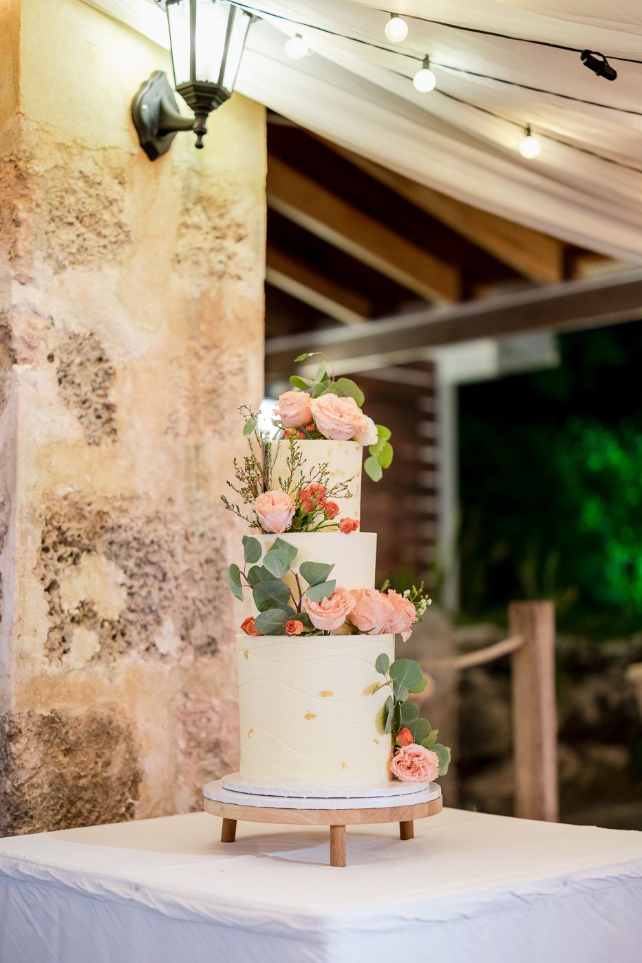 Three-tier white wedding cake adorned with peach roses and eucalyptus greenery on a wooden stand at an outdoor venue.