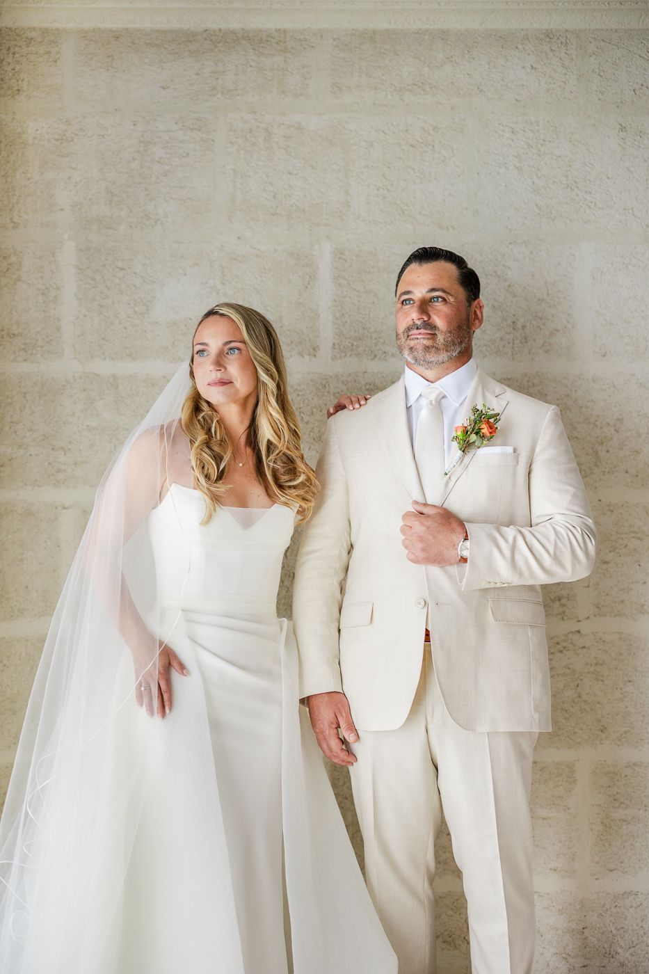 Bride in white gown with veil and groom in cream suit with floral boutonniere pose together against stone wall.