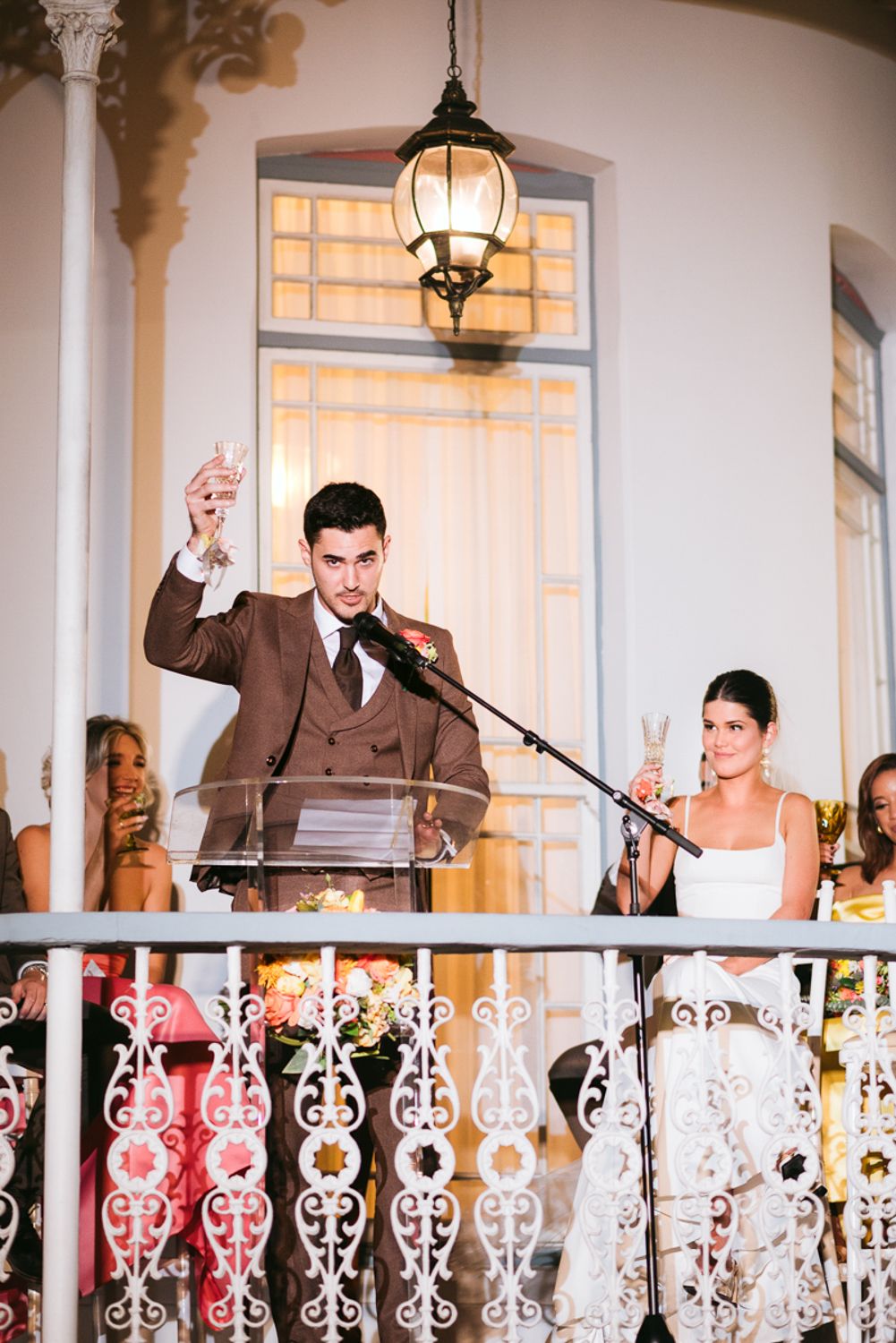 People give speeches during a wedding reception on an elegant porch with white wrought iron railings and hanging lanterns.