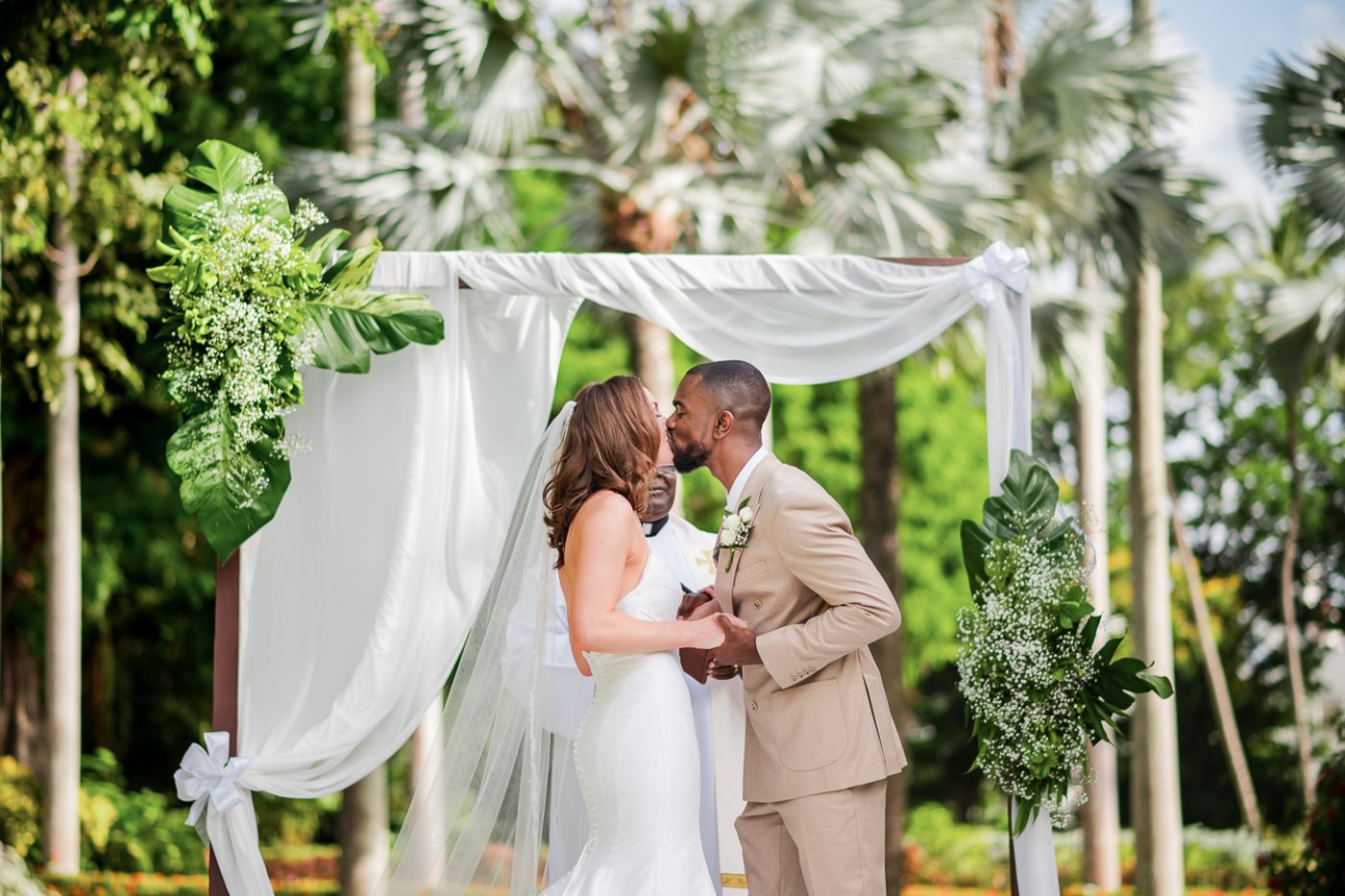 A romantic outdoor wedding ceremony taking place under white fabric draped between palm trees.