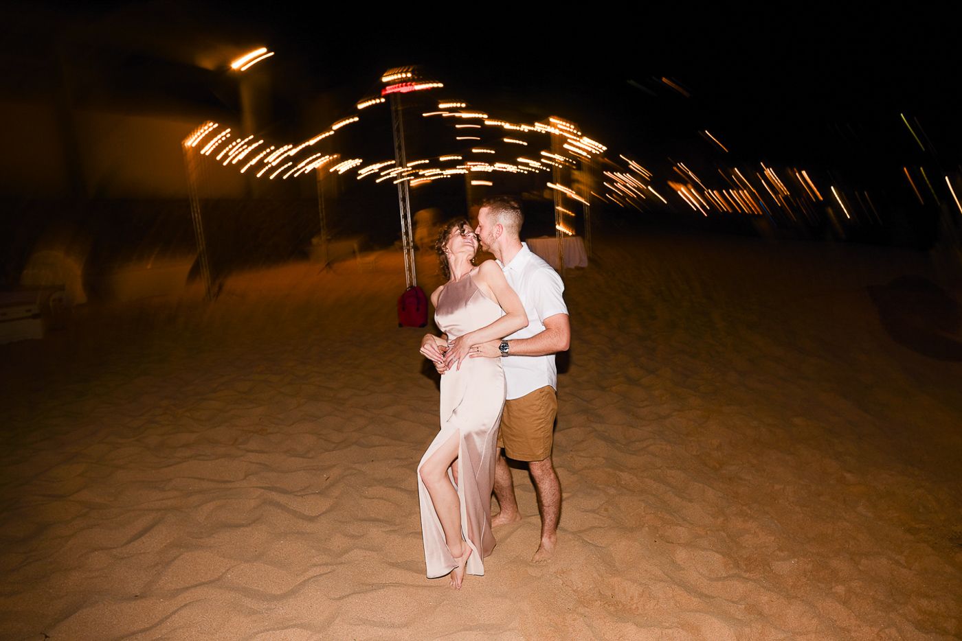Friends dance together at a beachside wedding reception under string lights at night.