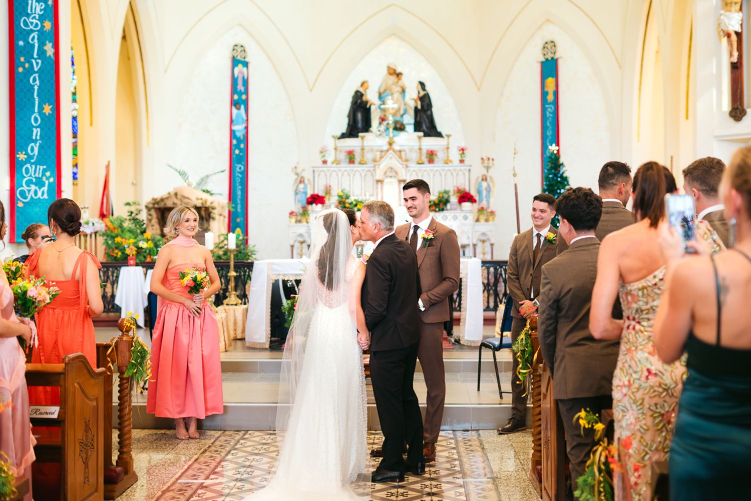 Guests gather in an elegant white ballroom for a wedding ceremony surrounded by floral arrangements and decorative columns.