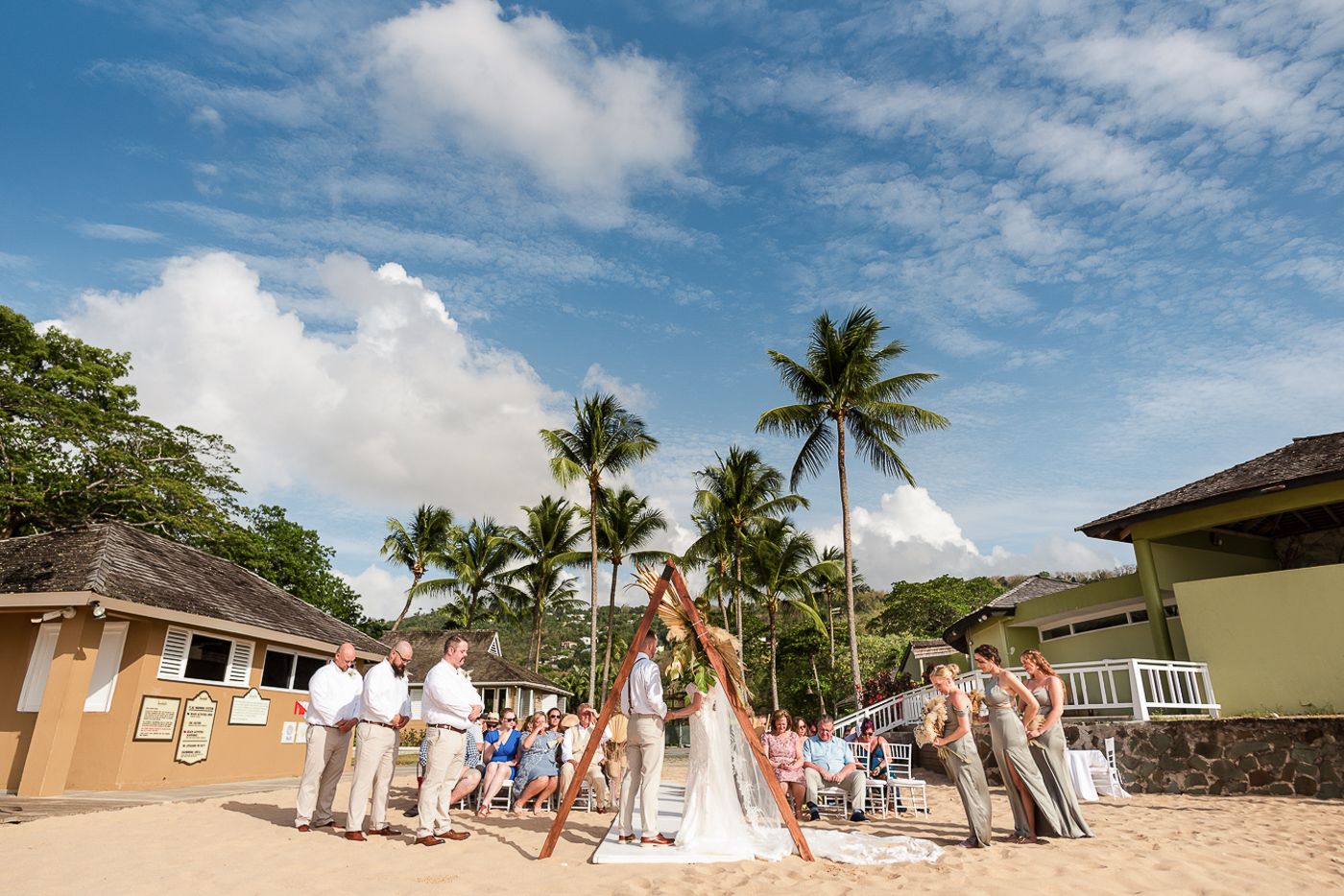 Tropical beach wedding venue with palm trees and yellow buildings in background.