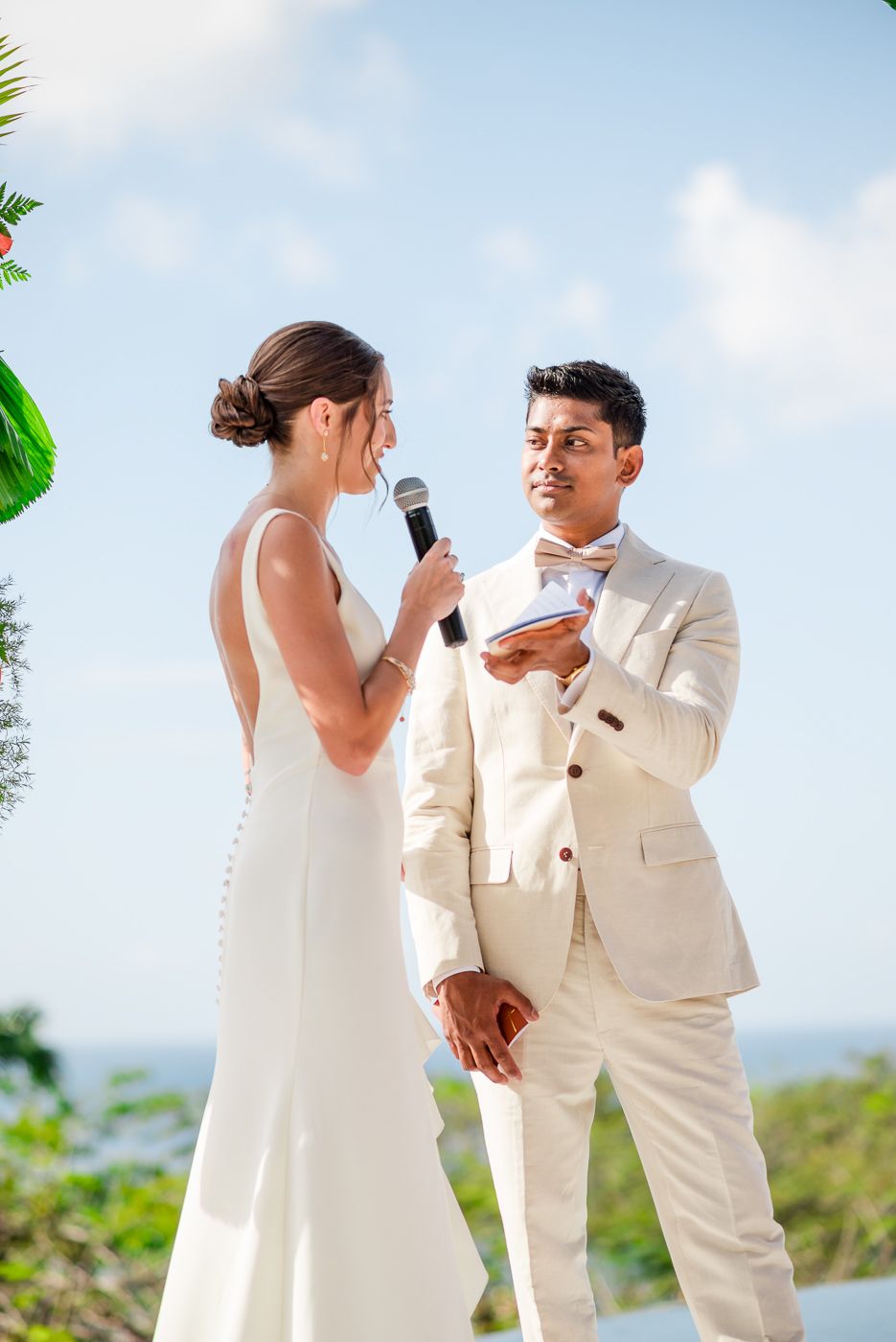 Wedding ceremony taking place outdoors with couple exchanging vows against tropical backdrop.
