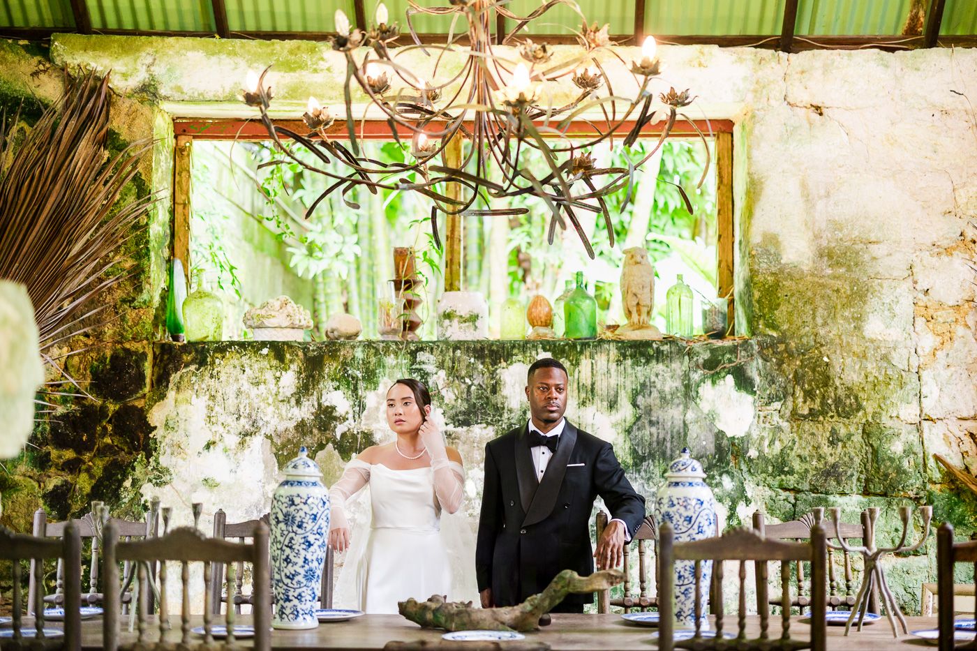 A rustic wedding reception table setup with an elegant chandelier and weathered walls in a greenhouse-style venue.