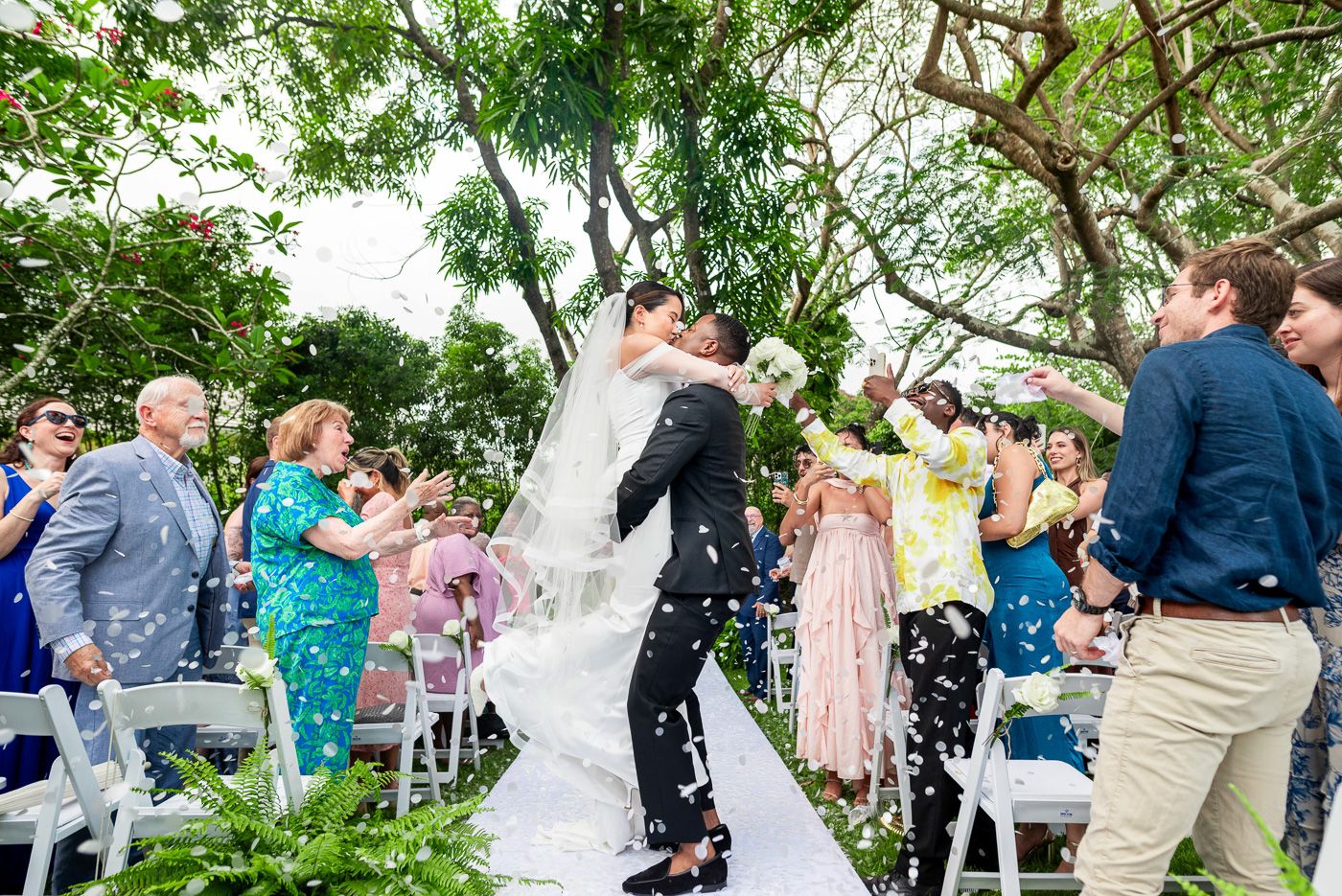 Wedding guests celebrate as the newlyweds share their first kiss under green trees.