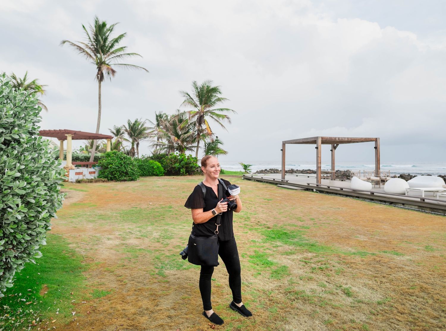 Woman in black outfit holding phone stands on tropical lawn with palm trees and ocean views on a cloudy day.