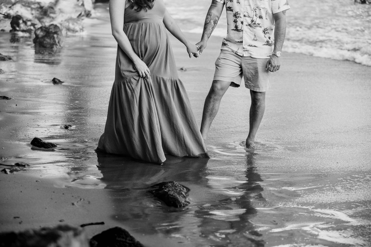 Black and white photo of a pregnant couple holding hands while walking along a sandy beach with ocean waves.