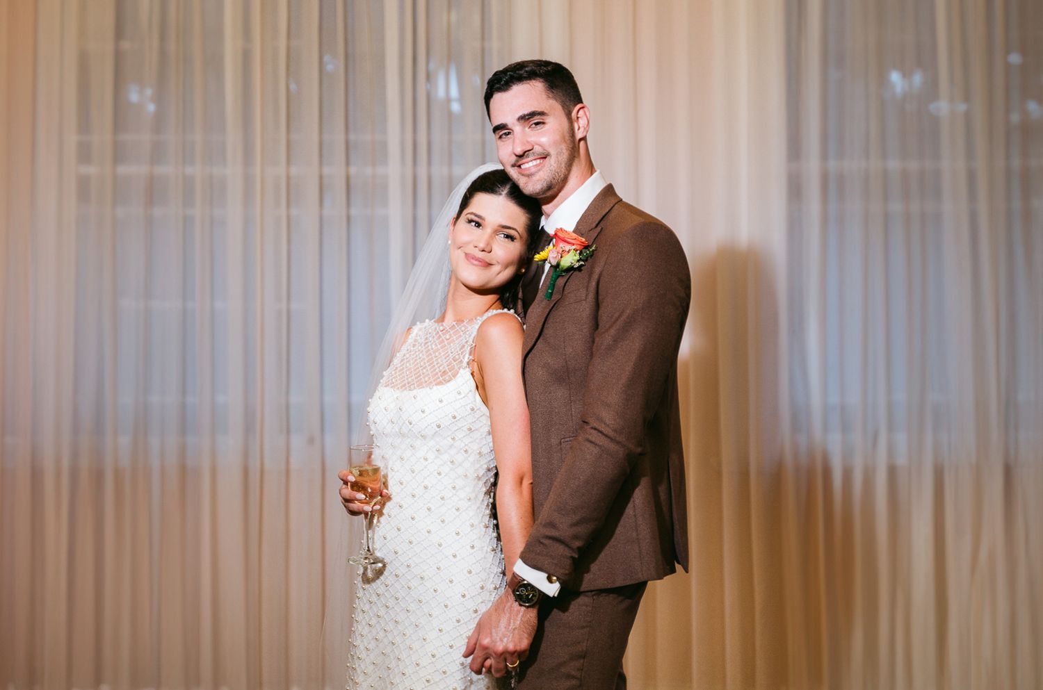 Bride and groom posing together in elegant wedding attire, she in white lace dress, he in brown suit with floral boutonniere.