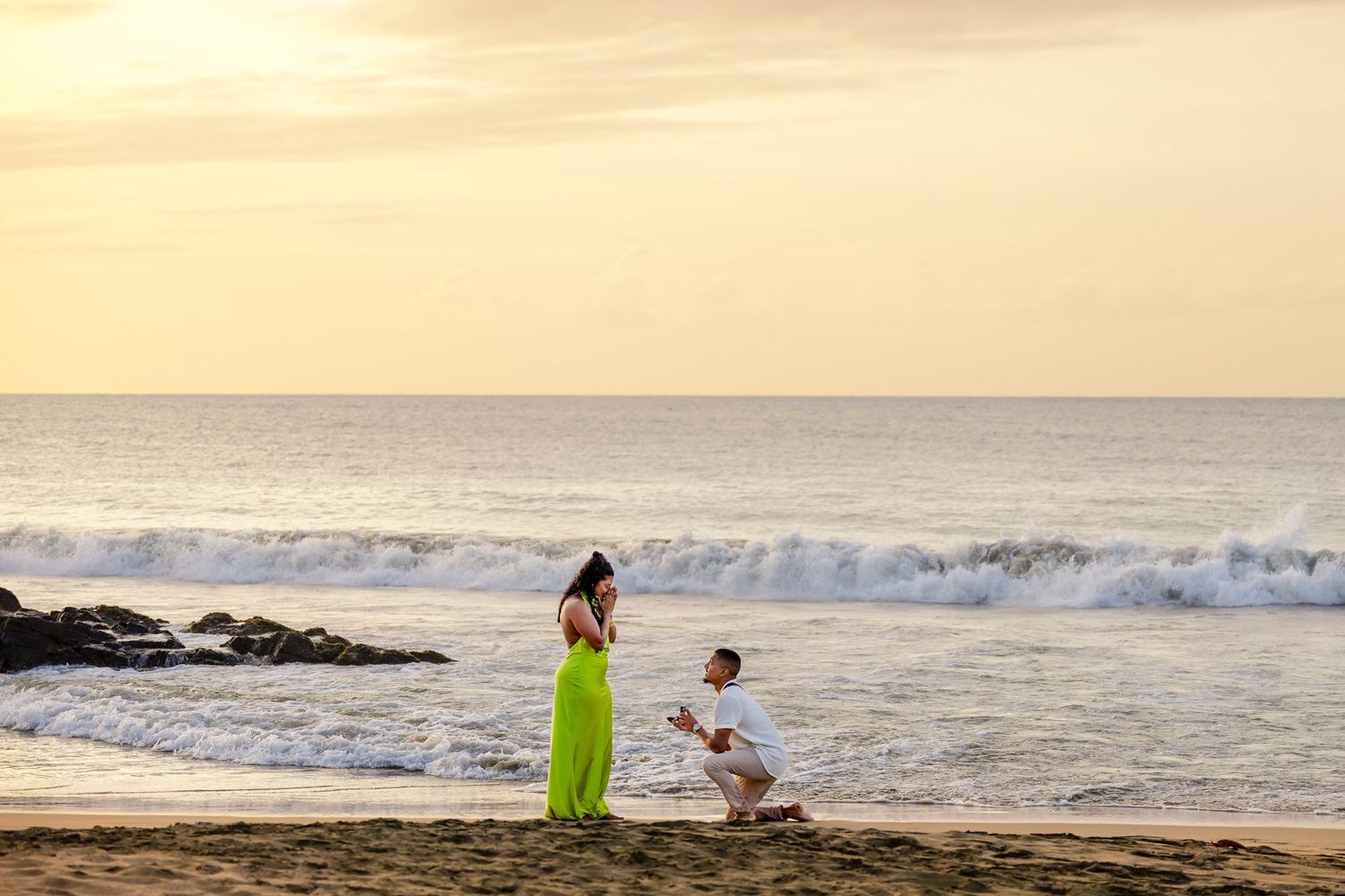 Man proposing on one knee to woman in green dress on sandy beach at sunset with ocean waves.