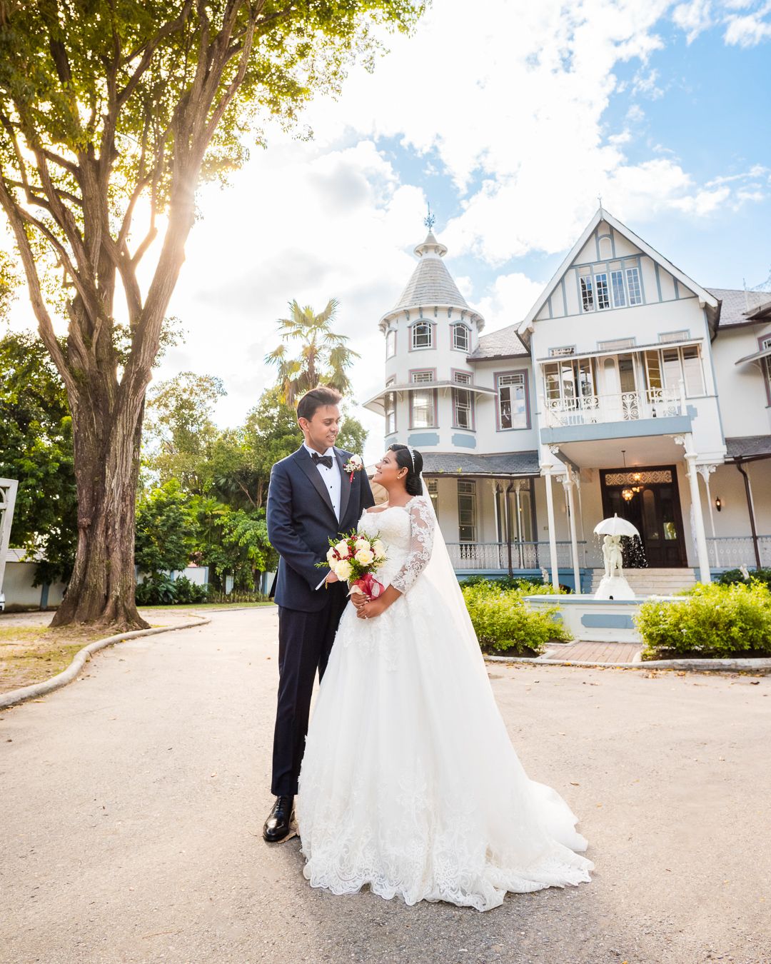Bride and groom stand together outside a Victorian mansion on their wedding day, surrounded by lush trees and blue skies.